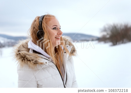 Woman in white down jacket and with headphones on her head smiling and walk on snowy winter day. Woman in white down jacket and with headphones on her head smiling and walk on snowy winter day. 108372218