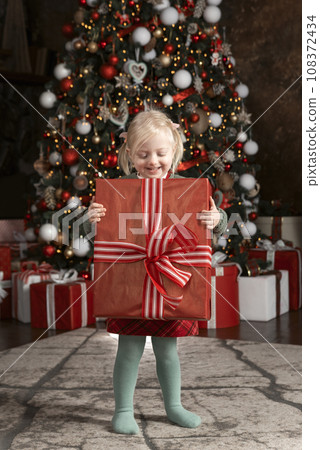 Smiling cute girl with big gift. Little blonde girl holds box with present, xmas tree background. Merry Christmas Smiling cute girl with big gift. Little blonde girl holds box with present, xmas tree background. Merry Christmas 108372434