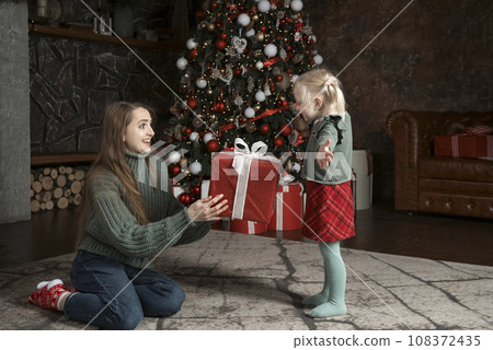 Small daughter gives big gift to her mom near Christmas tree. Portrait of mother and daughter on New Year Eve. Small daughter gives big gift to her mom near Christmas tree. Portrait of mother and daughter on New Year Eve. 108372435