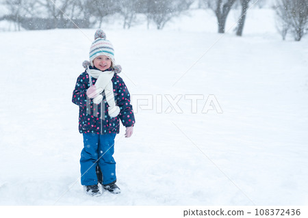 Full-length portrait of small child in knitted hat walks outside in winter during snowfall. Happy childhood. Winter holiday. Full-length portrait of small child in knitted hat walks outside in winter during snowfall. Happy childhood. Winter holiday. 108372436