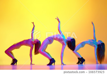 Flexibility. Beautiful three women dancing high heel dance, training over gradient yellow background in neon light 108373923