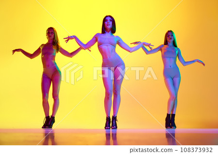 Slim, beautiful young women in shorts and top dancing in heels over gradient yellow background in neon light Slim, beautiful young women in shorts and top dancing in heels over gradient yellow background in neon light 108373932