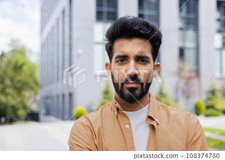 Close-up portrait of young Indian man, serious man concentrating and thoughtfully looking at camera, businessman outside office building. Close-up portrait of young Indian man, serious man concentrating and thoughtfully looking at camera, businessman outside office building. 108374780