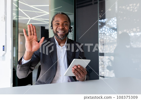 Help desk worker inside office at workplace, man smiling and looking at camera, waving greeting gesture, talking remotely, using headset phone and tablet for video call, consulting customer support. 108375309