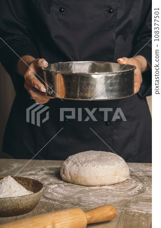 A woman sifts flour through a sieve over the dough close-up. The cook prepares bread dough 108377501