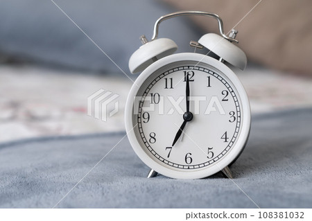 Close-up of a round white alarm clock on a table in the bedroom. The hands on the clock show seven o'clock in the morning, time to get up. Retro alarm clock on the table, vintage tone. space for text Close-up of a round white alarm clock on a table in the bedroom. The hands on the clock show seven o'clock in the morning, time to get up. Retro alarm clock on the table, vintage tone. space for text 108381032