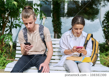Teenage students sitting on bench with backpacks, using smartphones Teenage students sitting on bench with backpacks, using smartphones 108381300