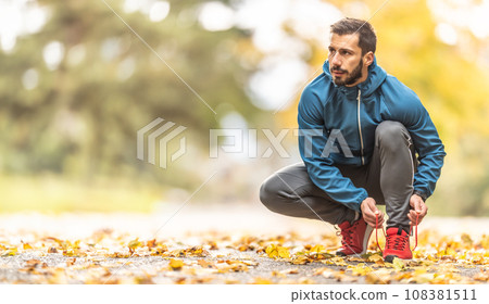 A young athlete in warm sportswear ties his shoelaces while running in an autumn park A young athlete in warm sportswear ties his shoelaces while running in an autumn park 108381511