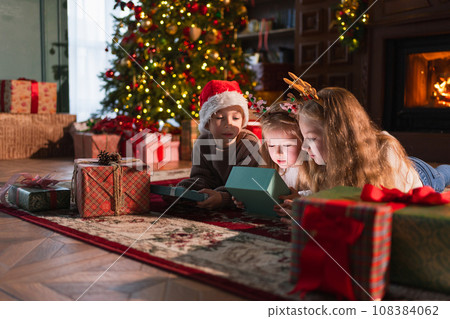 Merry Christmas Xmas holiday. Children unpacking open up gift box near Christmas tree. Kids in living room with Christmas tree fireplace opening gift box with surprise faces. Christmas eve at home Merry Christmas Xmas holiday. Children unpacking open up gift box near Christmas tree. Kids in living room with Christmas tree fireplace opening gift box with surprise faces. Christmas eve at home 108384062