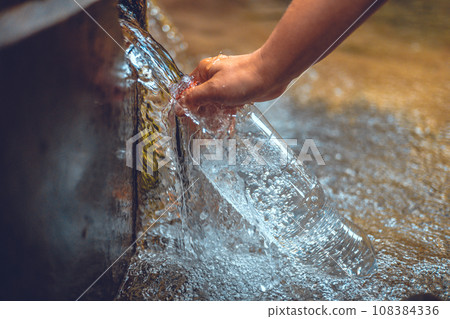 A traveler collects a bottle of cold, clean water from a spring. The man stopped to get clean water and drink from the spring. Clear, clean water flows in a stream into the offered bottle. 108384336