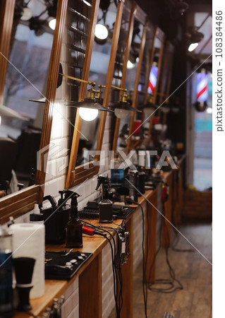 stylish barbershop with places for hairdresser artists, mirrors with illumination, work barber tools, razor, electric clippers, scissors, combs. interior with Wooden details and white tiles on wall 108384486