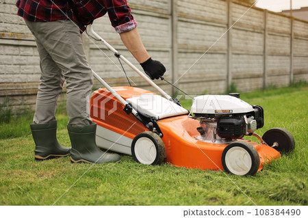 Unrecognisable gardener man in protective gloves starts the lawnmower before cutting green grass lawn in his backyard. Man with motorised lawnmower cares for landscaping lawn 108384490