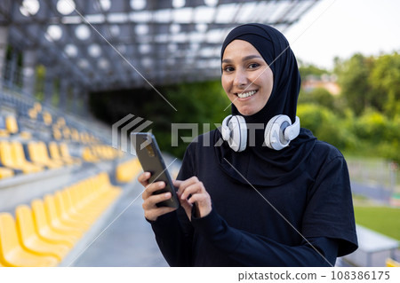 Portrait of a young hijab Muslim woman in a sports stadium running and doing active physical exercises, smiling and looking at the camera, using the phone and headphones to listen to music. 108386175