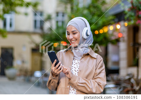 Young beautiful woman walking in the city, Muslim woman in hijab on evening walk in headphones listening to music smiling joyfully using smartphone app. 108386191