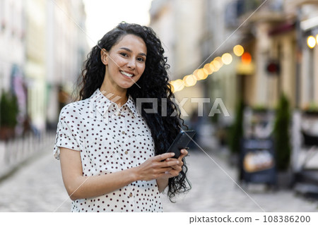 Portrait of a young beautiful Latin American woman walking in the evening city, holding a phone, smiling and looking at the camera. Portrait of a young beautiful Latin American woman walking in the evening city, holding a phone, smiling and looking at the camera. 108386200