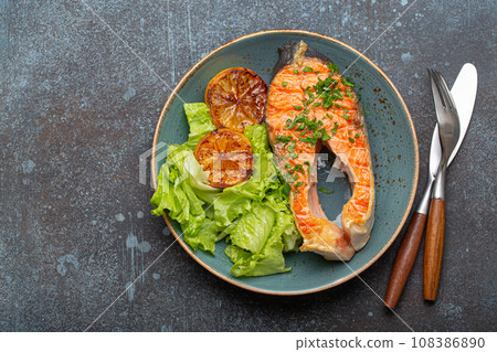 Grilled fish salmon steak and green salad with lemon on ceramic plate on rustic blue stone background top view, balanced diet or healthy nutrition meal with salmon and veggies, space for text Grilled fish salmon steak and green salad with lemon on ceramic plate on rustic blue stone background top view, balanced diet or healthy nutrition meal with salmon and veggies, space for text 108386890