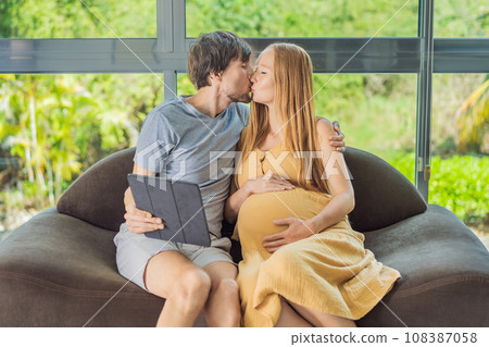 A smiling husband and his pregnant wife share a moment of joy, gazing at a tablet screen, their faces glowing with anticipation and happiness 108387058