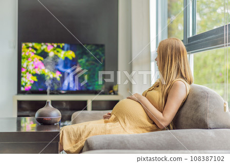 A blissful pregnant woman immerses in relaxation, savoring the soothing aroma from a diffuser while indulging in a calming TV video, embracing tranquility during her pregnant journey 108387102