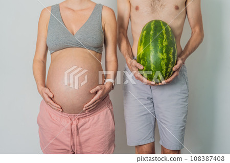 A humorous image: a pregnant woman and her husband playfully use a watermelon in place of a belly, comically highlighting the challenges of navigating with a pregnant bump 108387408