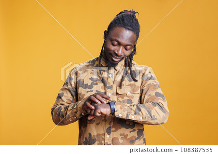 Portrait of businessman looking at wristwatch checking time, being late at work. African american entrepreneur worrying and being impatient about punctuality in studio over yellow background. Portrait of businessman looking at wristwatch checking time, being late at work. African american entrepreneur worrying and being impatient about punctuality in studio over yellow background. 108387535