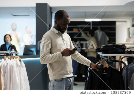 African american man scrolling clothing store online website on smartphone while shopping in department mall. Boutique customer checking shirt size in stock using mobile phone app African american man scrolling clothing store online website on smartphone while shopping in department mall. Boutique customer checking shirt size in stock using mobile phone app 108387569