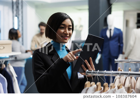 Smiling young asian woman analyzing apparel stock on digital tablet while working in retail market clothing store. Shopping mall employee managing garment merchandise inventory Smiling young asian woman analyzing apparel stock on digital tablet while working in retail market clothing store. Shopping mall employee managing garment merchandise inventory 108387604