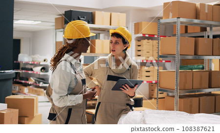 Two women with hardhats organizing products on shelves, looking at tablet to check stock logistics and planning distribution. Warehouse manager and employee counting cardboard packs. Two women with hardhats organizing products on shelves, looking at tablet to check stock logistics and planning distribution. Warehouse manager and employee counting cardboard packs. 108387621