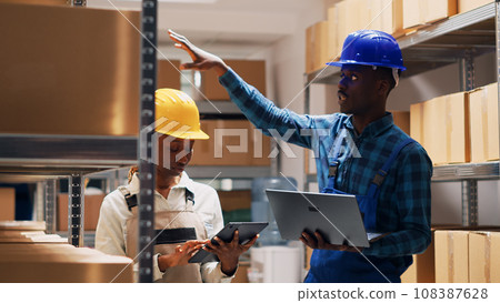 African american employees working on stock inventory, reading list of products on laptop and digital tablet. Man and woman checking merchandise on warehouse shelves, cargo goods in packs. 108387628