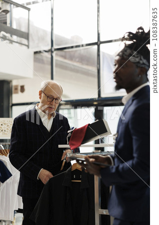 Senior client looking to buy stylish casual wear on hangers, checking shirt fabric in clothing shop. Customer wearing formal suit buying fashionable clothes and trendy accessories in modern boutique 108387635