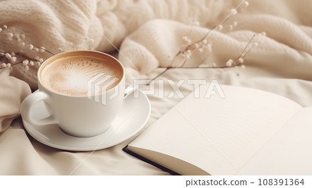 A cup of coffee with a book on a bed. The cup is white and has latte art. The book is white and open with blank pages. The bed has a white and beige blanket. White flowers are in the background. A 108391364