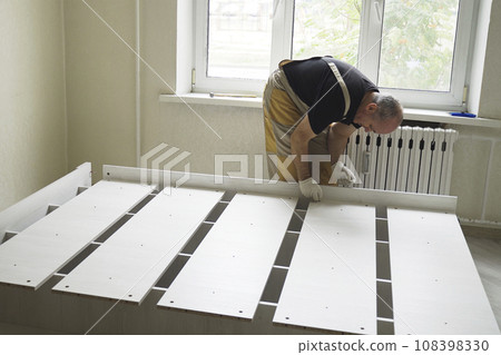 A male worker assembles furniture and connects the parts of the bed. A male worker assembles furniture and connects the parts of the bed. 108398330