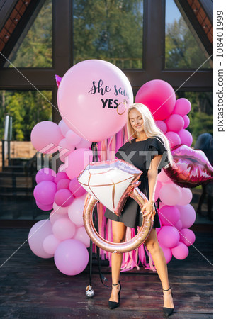 Vertical portrait of sensual blonde female in summer elegant dress posing holding various colorful festive foil helium balloons standing on background of country house building, sexy looking down. Vertical portrait of sensual blonde female in summer elegant dress posing holding various colorful festive foil helium balloons standing on background of country house building, sexy looking down. 108401999