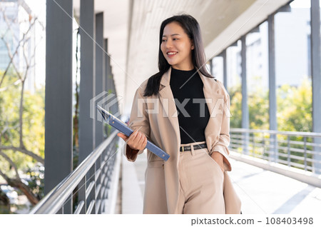 A beautiful Asian businesswoman is holding a document binder while walking on a skywalk. A beautiful Asian businesswoman is holding a document binder while walking on a skywalk. 108403498