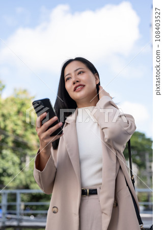 A beautiful Asian businesswoman stands outside in the city with her smartphone in her hand. 108403527