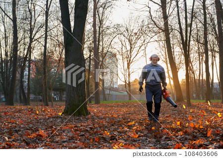 Serious senior man walking with handheld leaf blower to clean up park alley in October. Front view of focused bearded worker wearing uniform removing dry leaves in warm morning. Seasonal work concept. Serious senior man walking with handheld leaf blower to clean up park alley in October. Front view of focused bearded worker wearing uniform removing dry leaves in warm morning. Seasonal work concept. 108403606