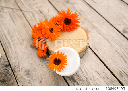 Cosmetic cream for cleansing the skin with calendula flowers. Medical Dermatology. Cream for body care with calendula. Fresh orange calendula flowers on a wooden background in nature. 108407495