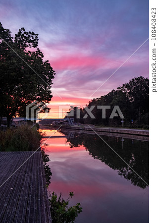 Dawn's Drama: Vibrant Sunrise Reflects in Canal, Framed by Tree Silhouettes Dawn's Drama: Vibrant Sunrise Reflects in Canal, Framed by Tree Silhouettes 108408423