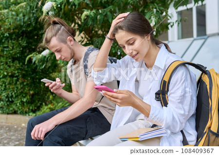 Teenage students sitting on bench with backpacks, using smartphones Teenage students sitting on bench with backpacks, using smartphones 108409367