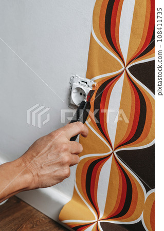 man cuts the edges of a wallpaper around a socket 108411735