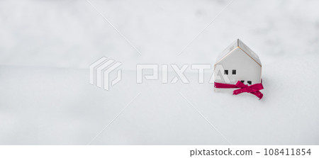 2024 Merry Christmas and happy new year horizontal banner with small toy model house wrapped in red satin ribbon on winter snow background. Miniature white toy house model  108411854