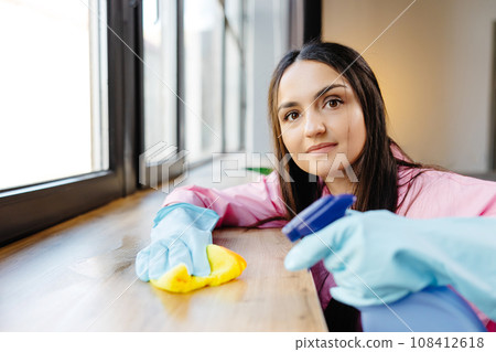 Woman in protective gloves is smiling and wiping dust using a spray and a duster while cleaning her house, close-up 108412618