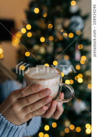 Closeup vertical cropped shot of unrecognizable little girl holding in hands mug with hot cocoa on blurred background bright bokeh lights of Christmas tree garland. Concept of home festive atmosphere. Closeup vertical cropped shot of unrecognizable little girl holding in hands mug with hot cocoa on blurred background bright bokeh lights of Christmas tree garland. Concept of home festive atmosphere. 108414240