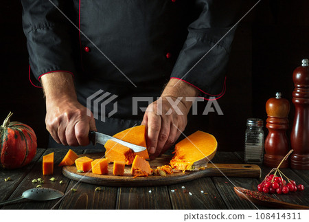 A man cuts a pumpkin with a kitchen knife on a wooden board. Fresh raw diced pumpkin on a kitchen cutting board. Preparing ingredients for a seasonal fall dish at home A man cuts a pumpkin with a kitchen knife on a wooden board. Fresh raw diced pumpkin on a kitchen cutting board. Preparing ingredients for a seasonal fall dish at home 108414311