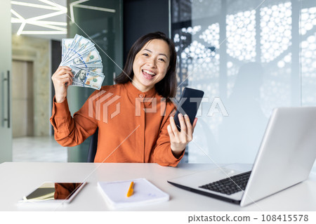 Portrait of young joyful asian business woman, woman smiling and looking at camera, holding phone and winning money cash american dollars, working at workplace inside office. Portrait of young joyful asian business woman, woman smiling and looking at camera, holding phone and winning money cash american dollars, working at workplace inside office. 108415578