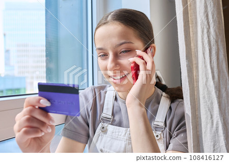 Teenage girl with credit bank card talking on cell phone at home 108416127