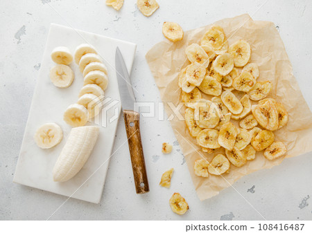 Fresh banana and baked crunchy chips snack on light kitchen background with knife.Top view. 108416487