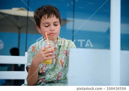 Hispanic handsome elementary age student boy in summer shirt, drinks fresh orange juice and studies online using laptop Hispanic handsome elementary age student boy in summer shirt, drinks fresh orange juice and studies online using laptop 108416599
