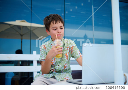 Confident smart elementary age student boy drinking juice and doing homework on laptop, sitting on a cafe summer terrace Confident smart elementary age student boy drinking juice and doing homework on laptop, sitting on a cafe summer terrace 108416600
