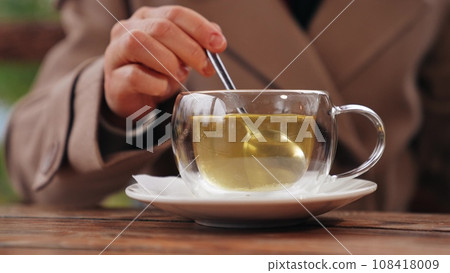 Woman stirs green tea with spoon in glass cup in cafe of park. Woman takes tea break with rotating spoon to stir sugar in cafe. Tea for maintaining woman figure with stirring spoon in cafe Woman stirs green tea with spoon in glass cup in cafe of park. Woman takes tea break with rotating spoon to stir sugar in cafe. Tea for maintaining woman figure with stirring spoon in cafe 108418009