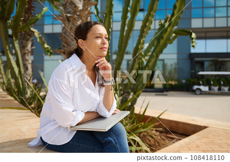 Beautiful woman in casual clothes, sitting on a stone bench with laptop on her knees, smiling and thoughtfully looking away 108418110